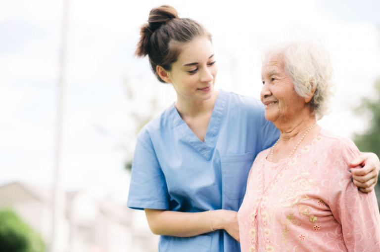 Caregiver in blue scrubs assisting elderly woman in pink blouse outdoors, emphasizing companionship and support in home care context.