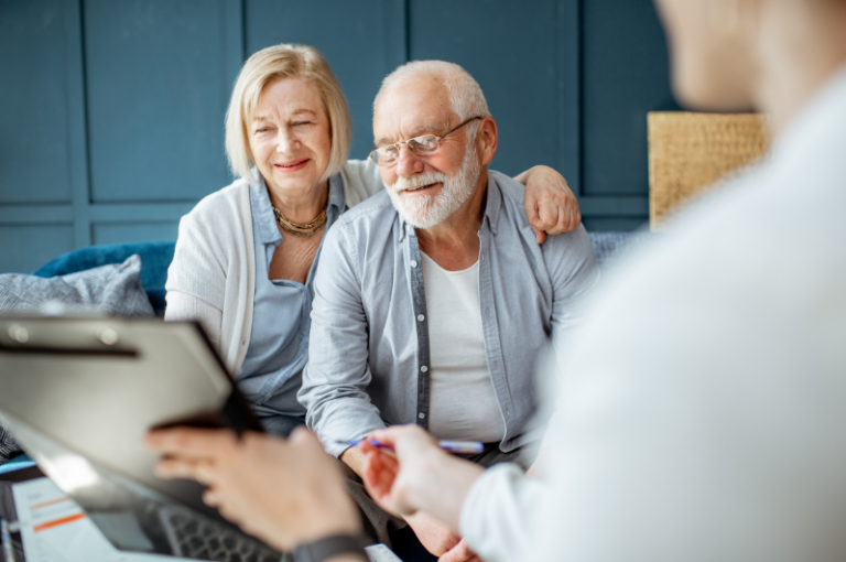 Elderly couple smiling and sitting together, engaging with a caregiver discussing end-of-life planning, with a clipboard and pen in focus.