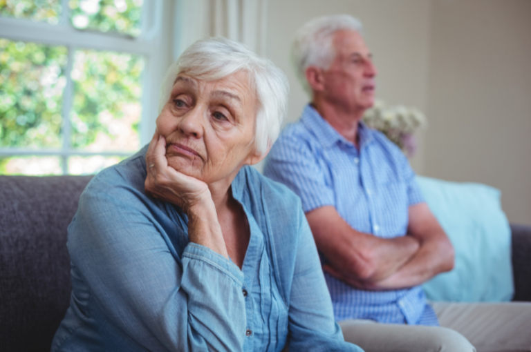 Elderly couple sitting apart on a sofa, with the woman looking contemplative and concerned, reflecting the emotional challenges in caregiving for a loved one with Alzheimer's.