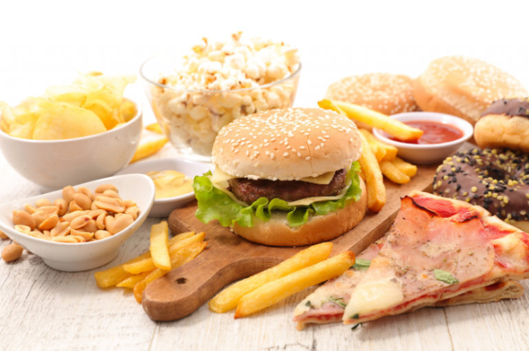 Assorted unhealthy foods including a sesame seed hamburger with lettuce, French fries, popcorn, potato chips, peanuts, pizza slice, and donuts on a wooden board.