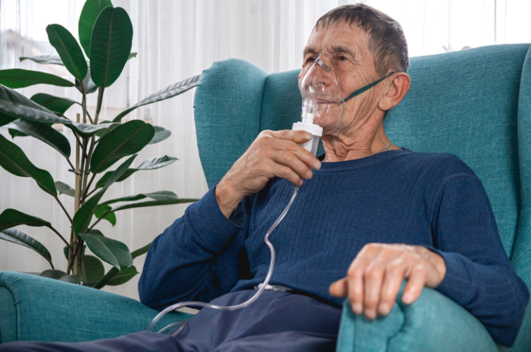 Elderly man using a nebulizer for respiratory treatment while seated in a comfortable chair, surrounded by plants, highlighting senior health and wellness.