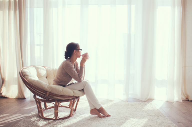 Woman sitting in a rattan chair, sipping a drink, reflecting in a sunlit room with sheer curtains, conveying relaxation and self-care in a home environment.