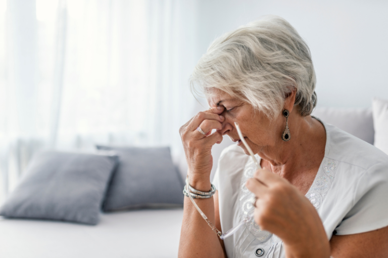 Elderly woman experiencing discomfort, holding her glasses, in a softly lit room, reflecting the challenges faced by seniors with cognitive issues and pain management.