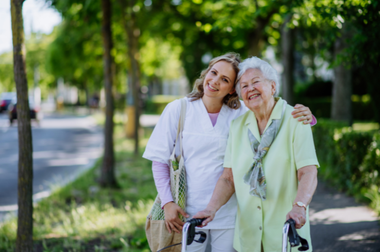 Caregiver assisting elderly woman with walker, smiling together in a green outdoor setting, representing companionship and support in senior care.