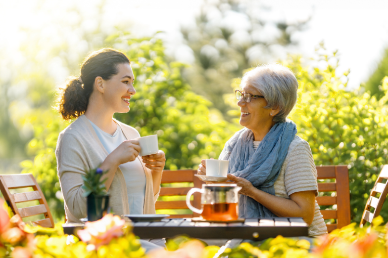 Two women enjoying tea outdoors, one younger with dark hair and a light sweater, and an older woman with gray hair and glasses, smiling while engaging in conversation amidst a lush garden setting, reflecting themes of senior care and companionship.
