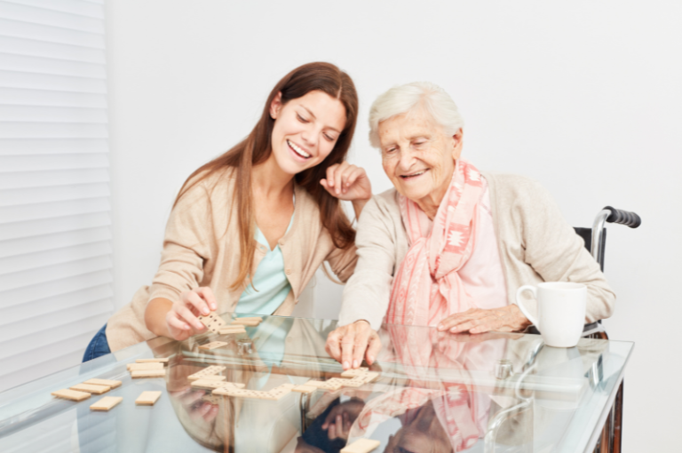 Young woman and elderly woman playing a game with wooden blocks at a glass table, promoting bonding activities for seniors and families.
