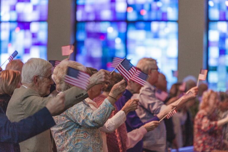 Elderly individuals waving small American flags during a ceremony, with colorful stained glass windows in the background, highlighting community and patriotism in a senior care setting.