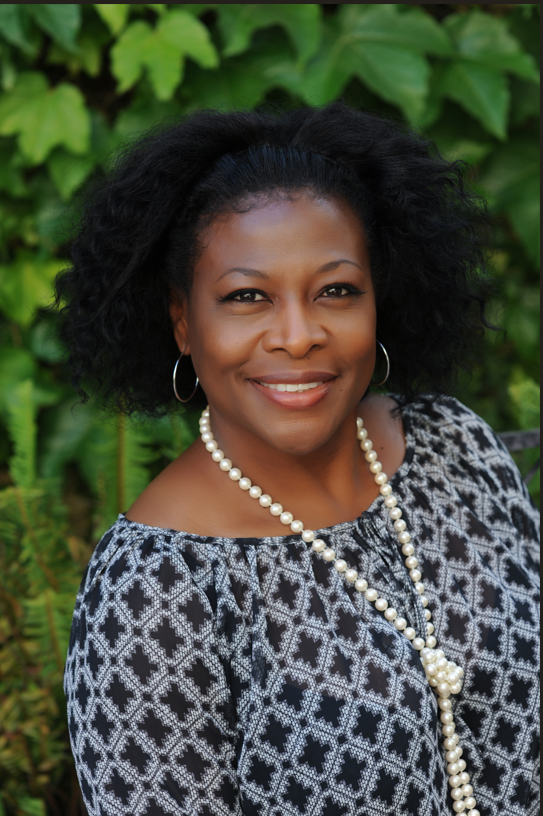 Smiling woman with curly hair wearing a patterned blouse and pearl necklace, standing against a backdrop of green foliage, representing compassionate in-home care services.